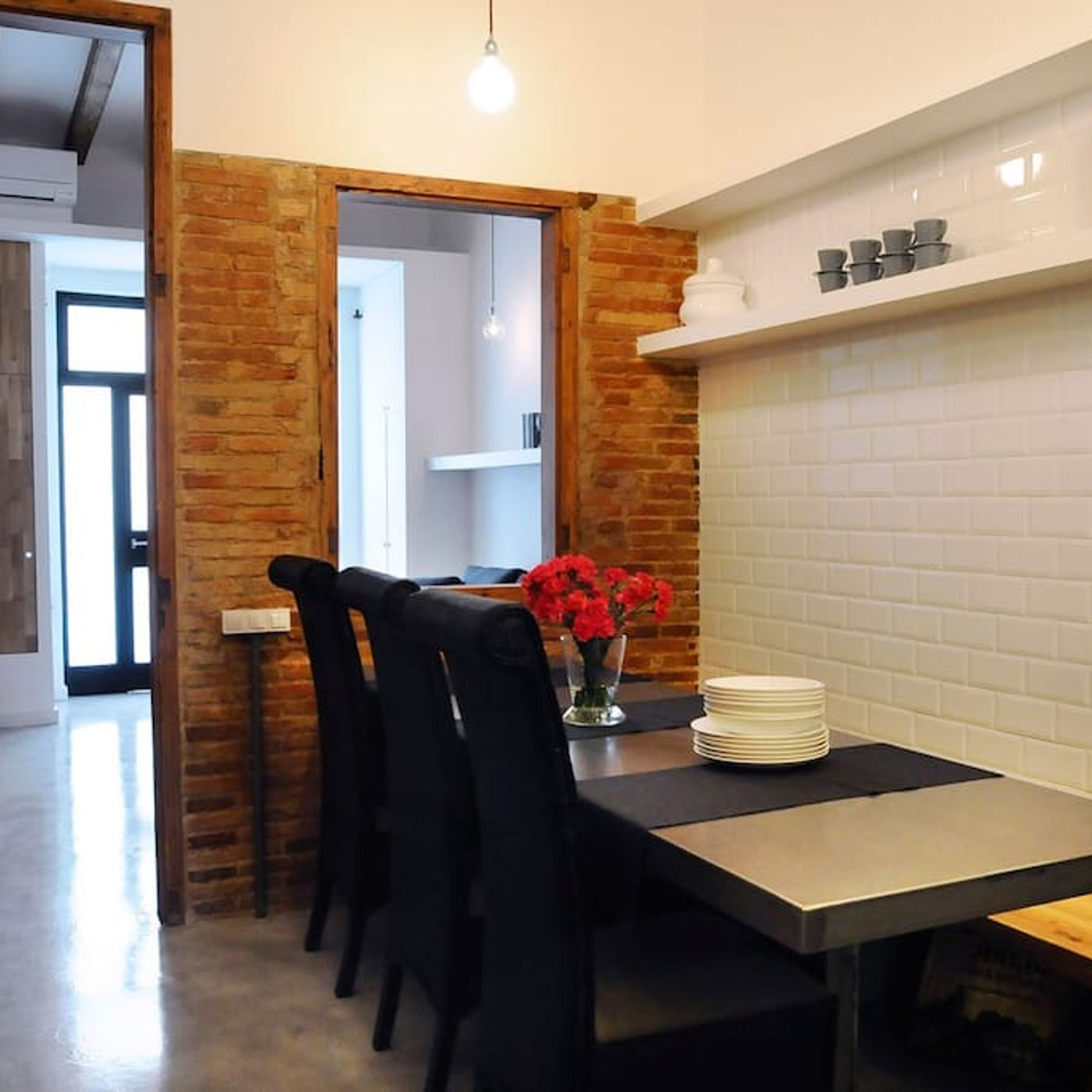 Un coin repas avec un mur en briques, un pare-feu en carreaux blancs et une table dressée avec des assiettes et un vase rempli de fleurs rouges.