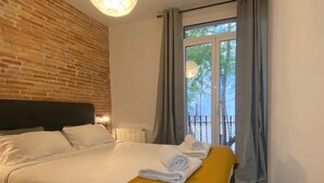 A hotel room with a brick wall, a bed with white linens, a yellow throw blanket, and a view of trees through the window.
