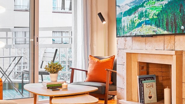 A modern living room with a fireplace, a television mounted on the wall, and a balcony with a view of buildings. - Love Local Life Near Sagrada Familia By Myrentalhost (Barcelona)