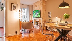 A modern living room with a wooden floor, a round dining table with chairs, a flat-screen TV mounted on the wall, and a fireplace. - Love Local Life Near Sagrada Familia By Myrentalhost (Barcelona)