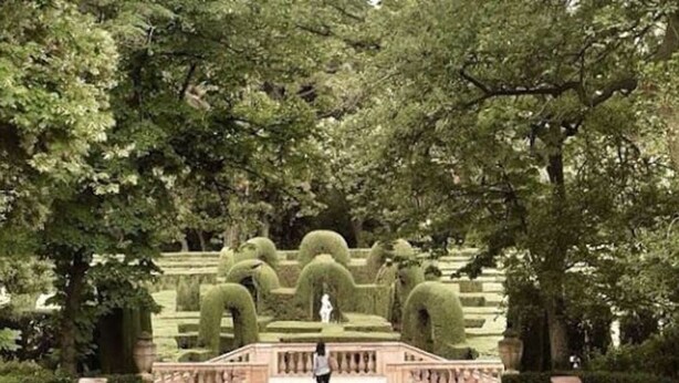 Ein gepflegter formaler Garten mit ordentlich beschnittenen Hecken, einer zentralen Statue und einer Person in der Mitte.