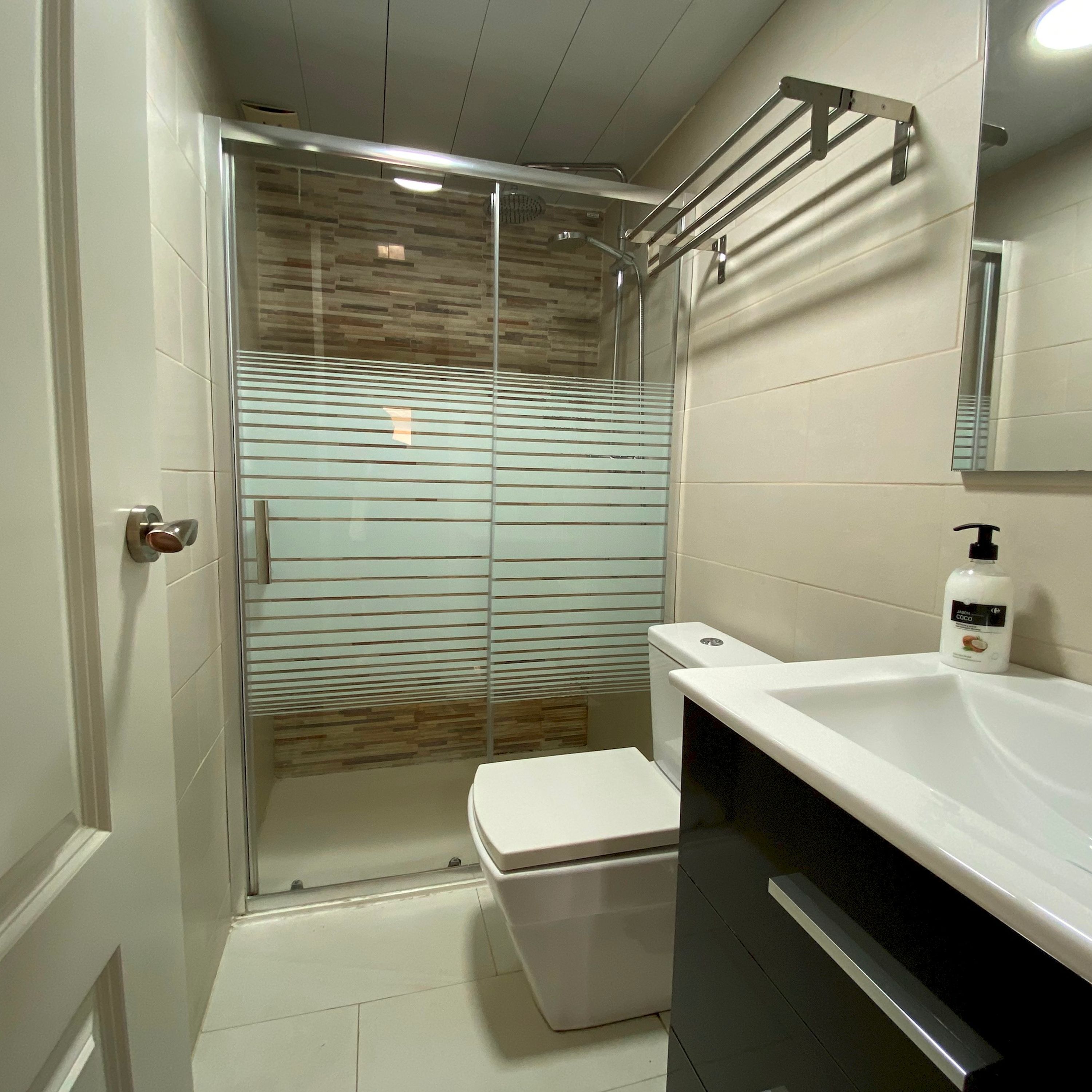 Ein Badezimmer mit Duschkabine aus Glas, einer weißen Toilette, einem Waschbecken mit Seifenspender und einem Spiegel an der Wand.