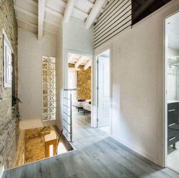 A narrow hallway with a wooden floor, a staircase, and a bed in the background.
