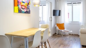 Een moderne woonkamer met een houten eettafel, witte stoelen, een kleurrijk abstract schilderij en een aan de muur bevestigde televisie.