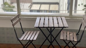 Une petite table pliante, accompagnée de deux chaises en bois, est placée devant une fenêtre donnant sur des immeubles.