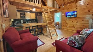 A cozy cabin interior with a wooden staircase, a kitchen area, a living room with red sofas, and a television mounted on the wall. - Pluggy’s Place - Perfect Location |Outdoor Firepit (Beattyville)