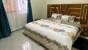 A neatly made bed with a patterned bedspread and pillows, a wooden headboard, and a window with curtains. - Elite Homes (Kampala)