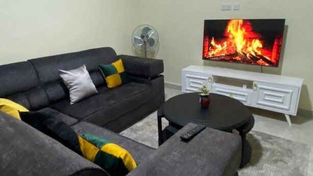 A living room with a sectional sofa, a flat-screen TV displaying a fire, a white sideboard, and a small table with a remote control. - Elite Homes (Kampala)