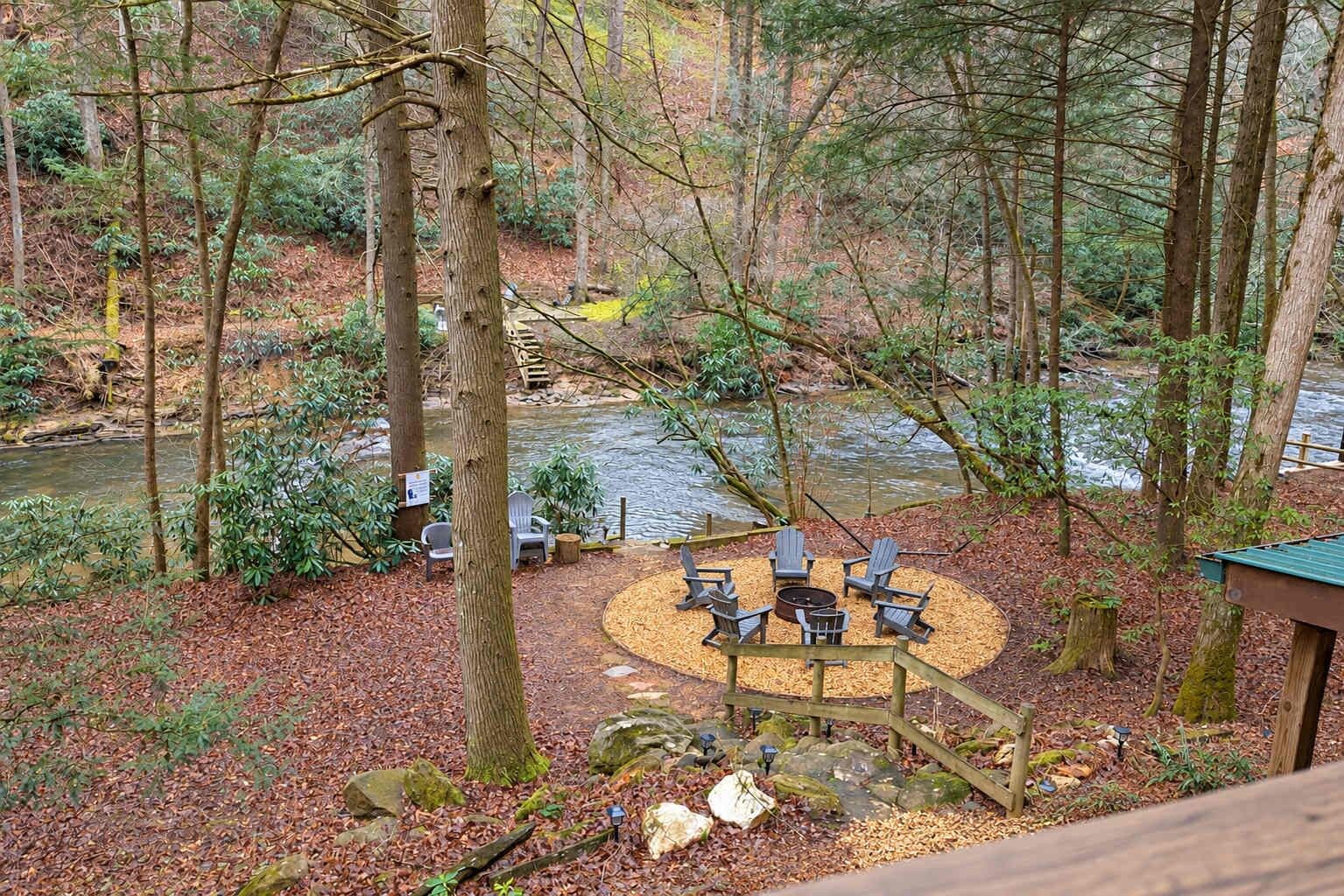 A circular outdoor seating area with chairs and a fire pit surrounded by a wooden deck, overlooking a river in a forested area.