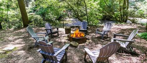 A circular seating arrangement around a campfire in a forested area with Adirondack chairs.
