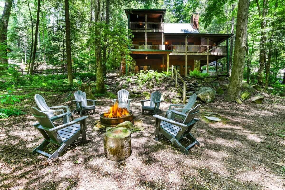 Mökki, jossa on terassi ja se on metsän ympäröimä. Siellä on Adirondack-tyylisiä puutarhatuoleja ja nuotiopaikka.