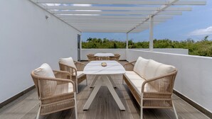 Un patio exterior con una mesa blanca y sillas de mimbre, una pérgola sobre la cabeza y vistas de vegetación al fondo.