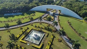 Vista aérea de un área ajardinada con una gran estructura curva, una plaza central con una fuente y palmeras.