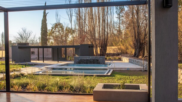 A modern outdoor pool area with a wooden deck, a stone structure, and a view of trees and a building in the background. - Amerian Chacras de Coria (Chacras de Coria)