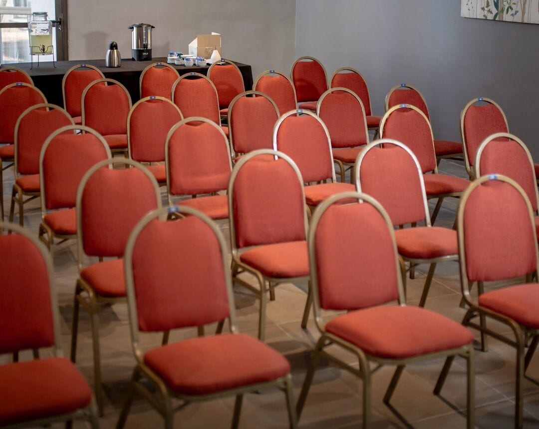 A conference room with rows of red chairs and a table with a coffee pot and other items.