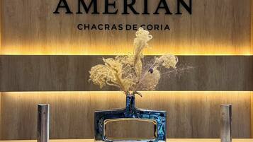 Un mostrador de recepción con un panel de madera de fondo, un letrero que dice 'AMÉRIAN CHACRAS DE CORIA' y un jarrón con flores secas.
