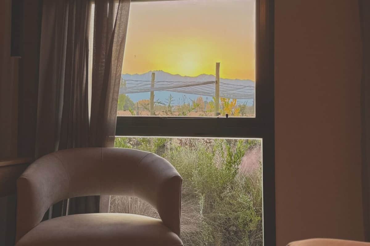 A room with a bed, a chair, and a window with a view of a sunset and a bridge.
