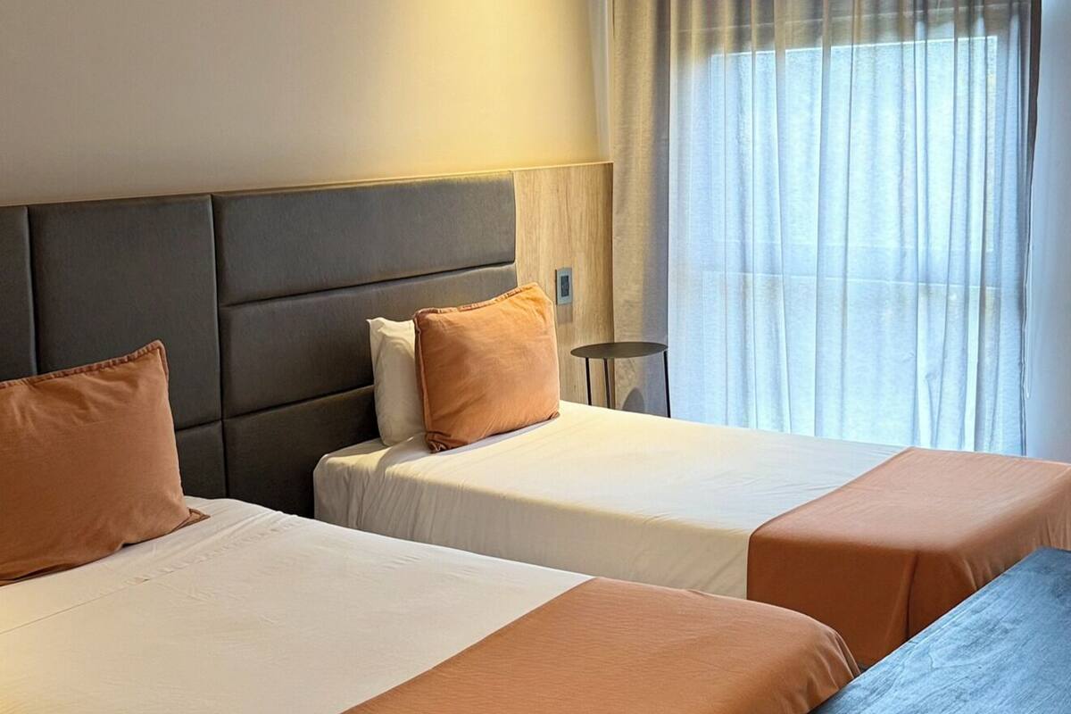 A hotel room with two beds, a bench, a bedside table, and a window with curtains.