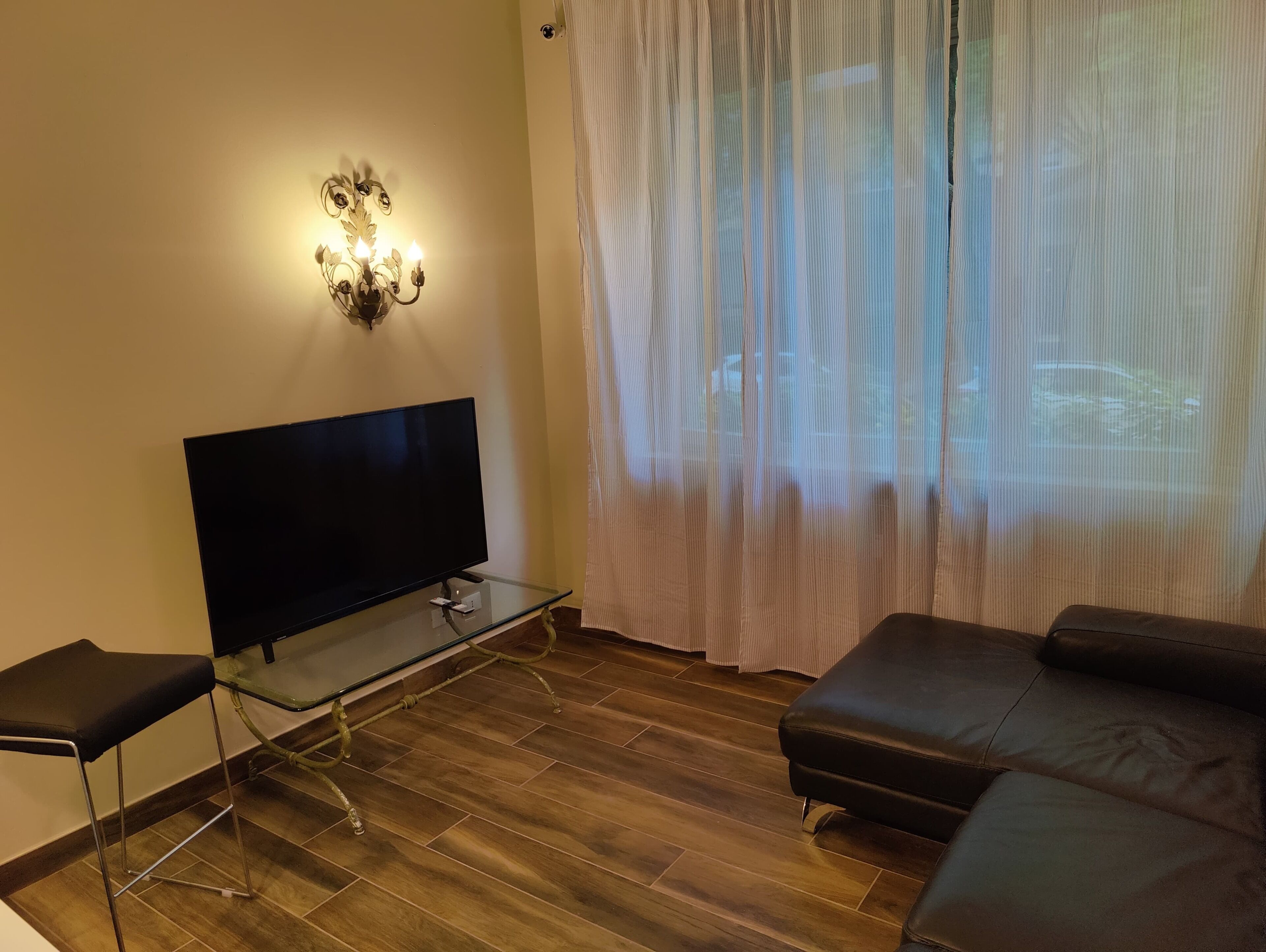 Ein Wohnzimmer mit Flachbildfernseher auf einem Glastisch, einer schwarzen Ledersofa und einem weißen Vorhang.