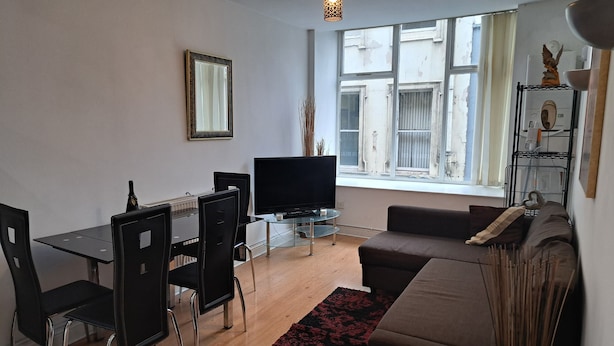 A modern living room with a glass dining table, black chairs, a brown sofa, a TV, and a large window with a view of buildings. - City Center Gem (Liverpool)