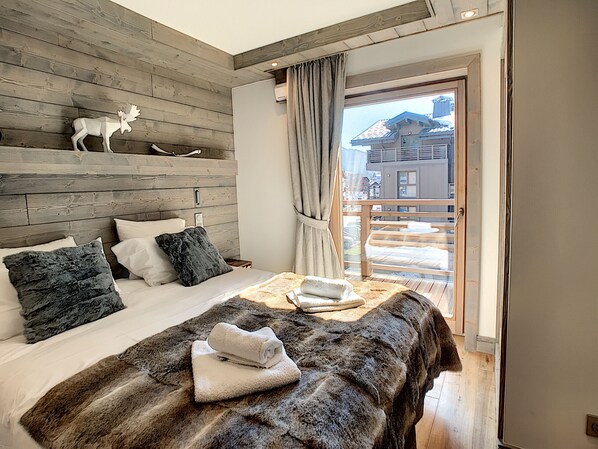 Un dormitorio con cabecera de madera, cama con una manta de piel y una toalla, mesita de noche de madera y vista a un edificio a través de una ventana.