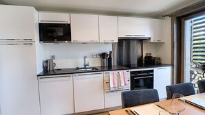A modern kitchen with white cabinets, a stainless steel range hood, and a built-in oven. There is a dining area with a wooden table and chairs. - Annapurna-appartement-6pers-proche centre (Les Gets)