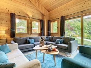 A cozy cabin interior with a sectional sofa, a small table with a kettle, and large windows offering a view of the surrounding greenery.