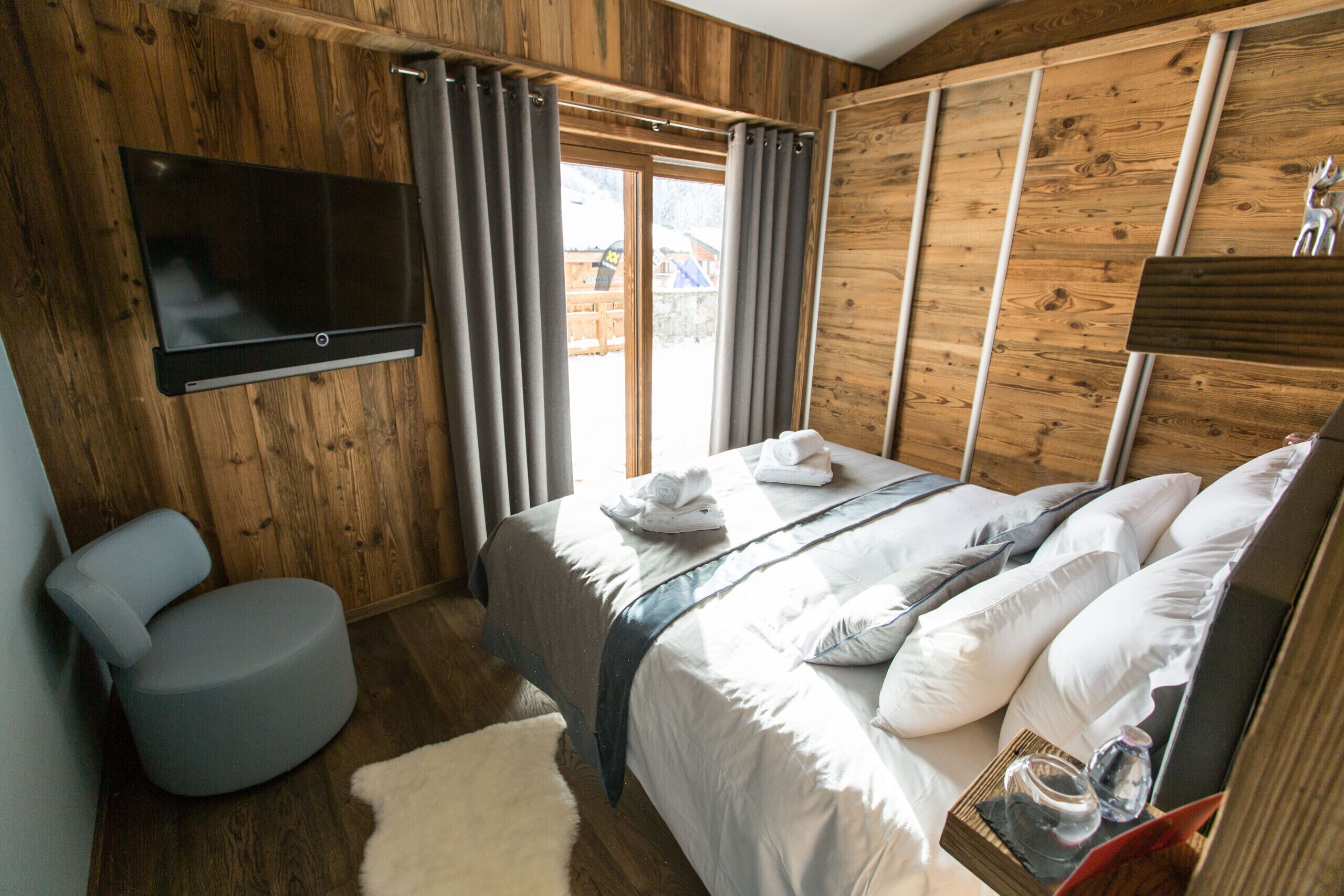 A bedroom with a bed, a TV mounted on the wall, and a wooden ceiling.