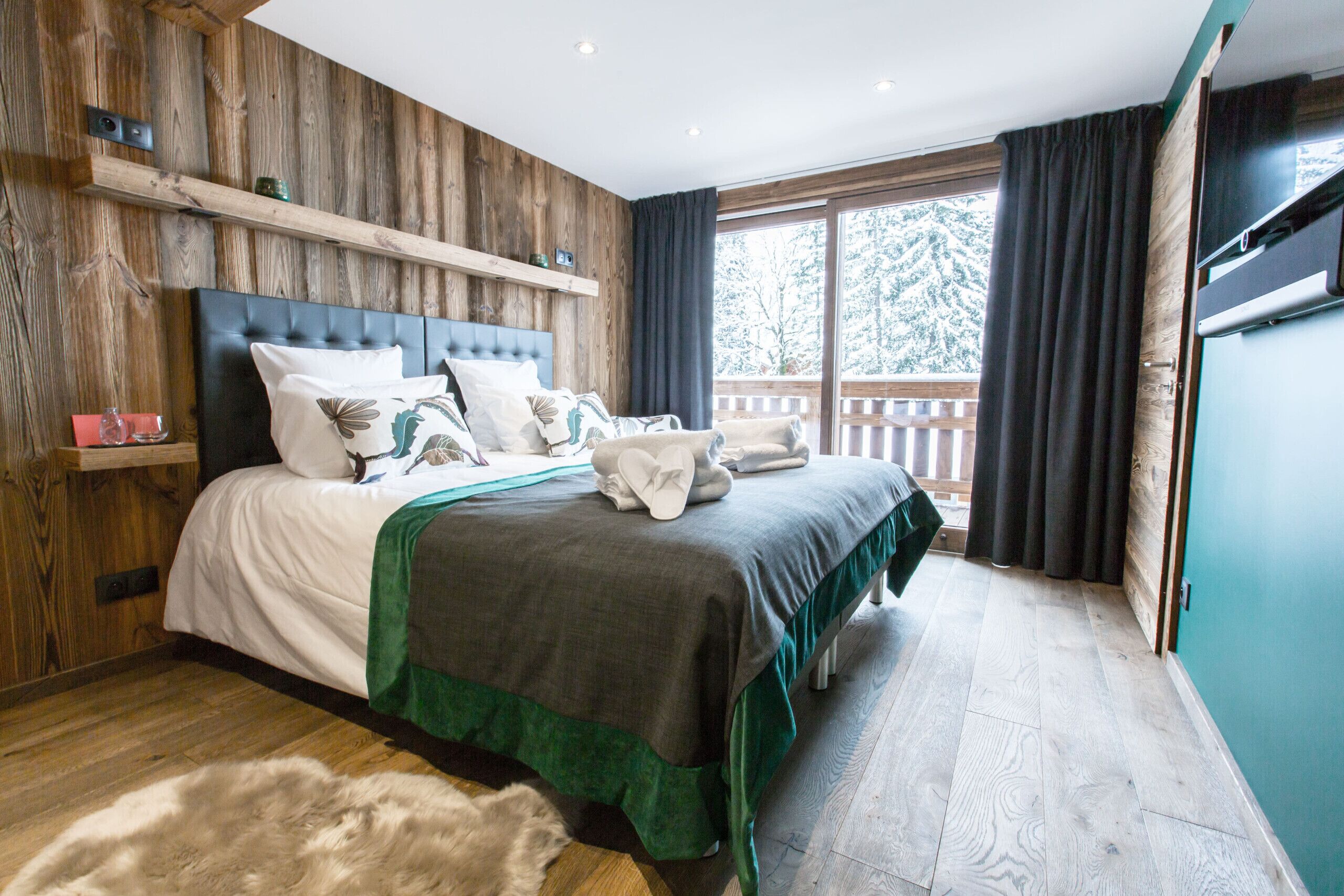 Ein Schlafzimmer mit einem hölzernen Kopfende, einem großen Bett mit vielen Kissen, einer grünen Tagesdecke und Blick auf eine Holzterrasse und Bäume durch ein Fenster.