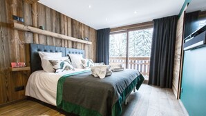 A bedroom with a wooden headboard, a large bed with multiple pillows, a green throw blanket, and a view of a wooden deck and trees through a window. - Chalet Le Saint-Jean - 15 pers - proche pistes (Les Gets)