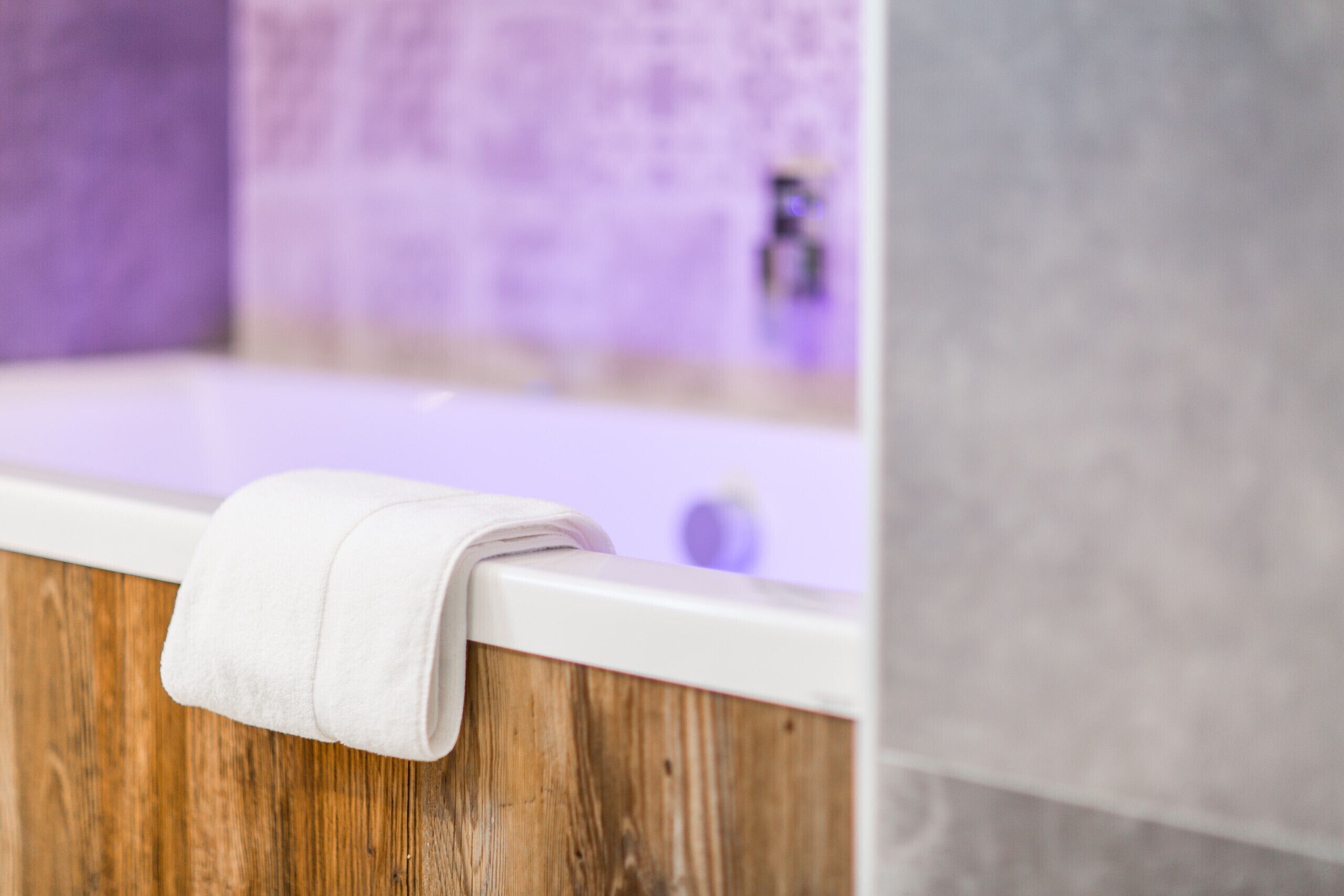 A bathtub with a white towel on the edge, wooden paneling, and a purple-tiled wall in the background.
