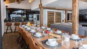 A modern dining area with a wooden table set for a meal, a kitchen in the background, and a cozy living space with a fireplace. - Chalet Le Saint-Jean - 15 pers - proche pistes (Les Gets)