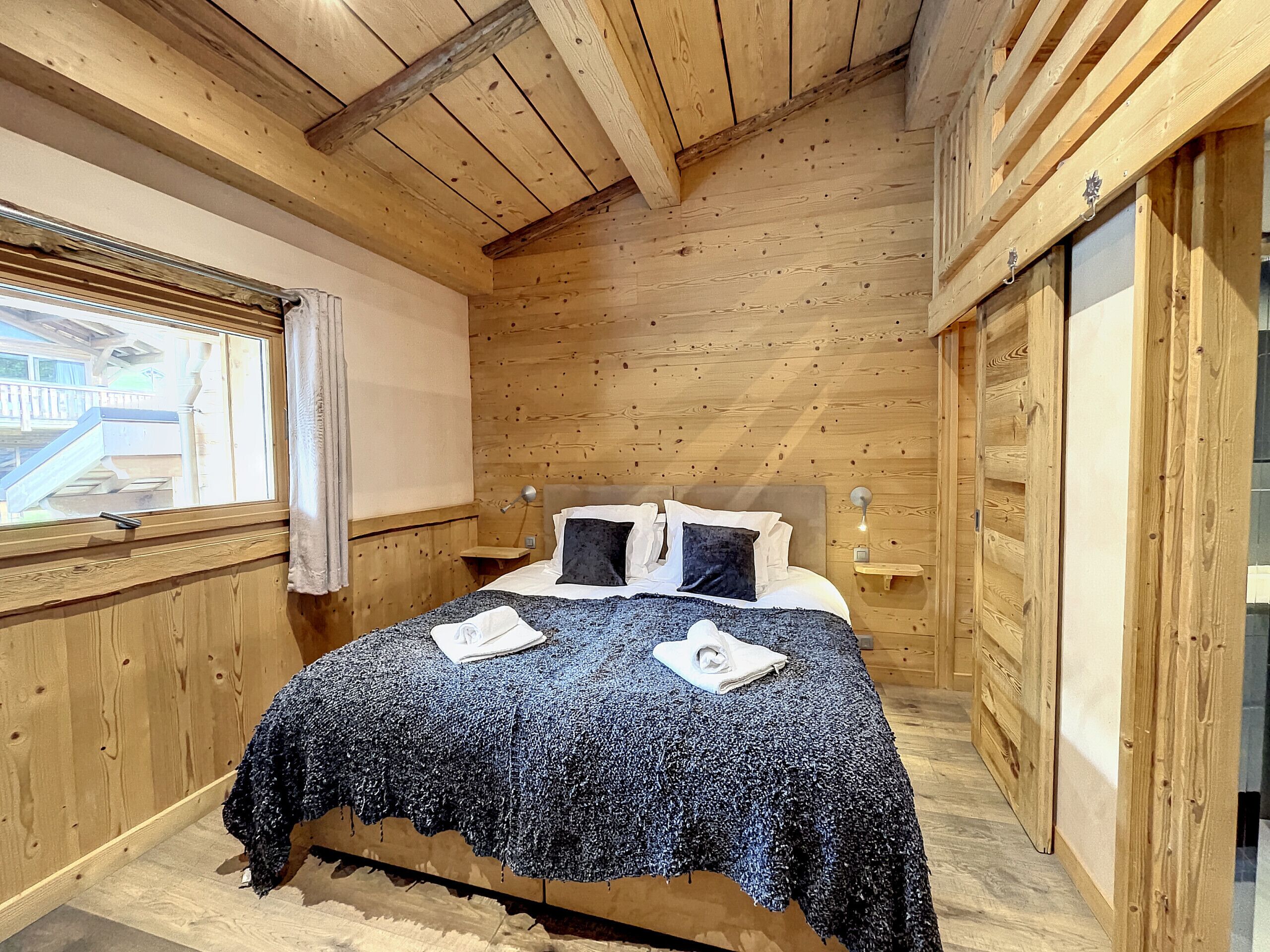 A wooden-paneled bedroom with a bed, a nightstand, a window, and a view of the outside.