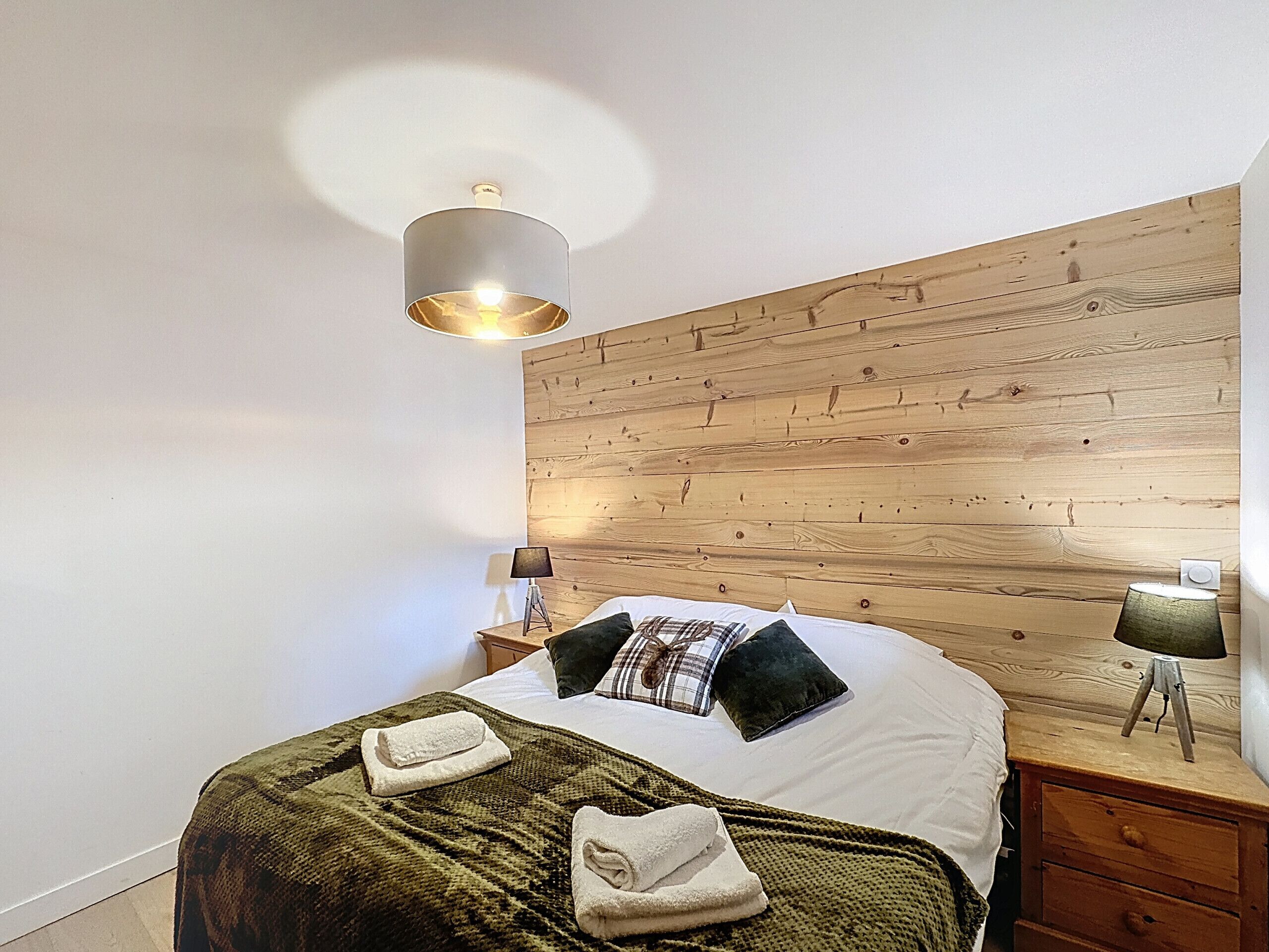 A bedroom with a wooden headboard, a bed with white linens, two pillows, a green blanket, and two bedside lamps.