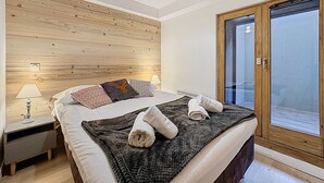 A modern bedroom with a wooden headboard, a large bed with multiple pillows, a bedside table with a lamp, and a glass door leading to a balcony. - La Bruyère - appartement - 6pers (Les Gets)