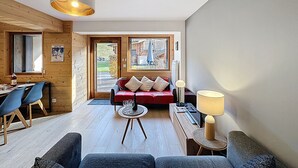 A modern living room with a wooden interior, a red sofa, a dining area with blue chairs, and a large window providing a view of the outside. - La Bruyère - appartement - 6pers (Les Gets)