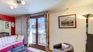 A room with a bed, a chair, a table, a TV, and a balcony with a view of buildings. - Clos 11 - appartement - 8 pers - proche centre (Les Gets)