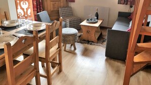 A cozy room with a wooden dining table set for four, a sofa, a small round table with a bottle and glasses, and a television on a wooden cabinet.