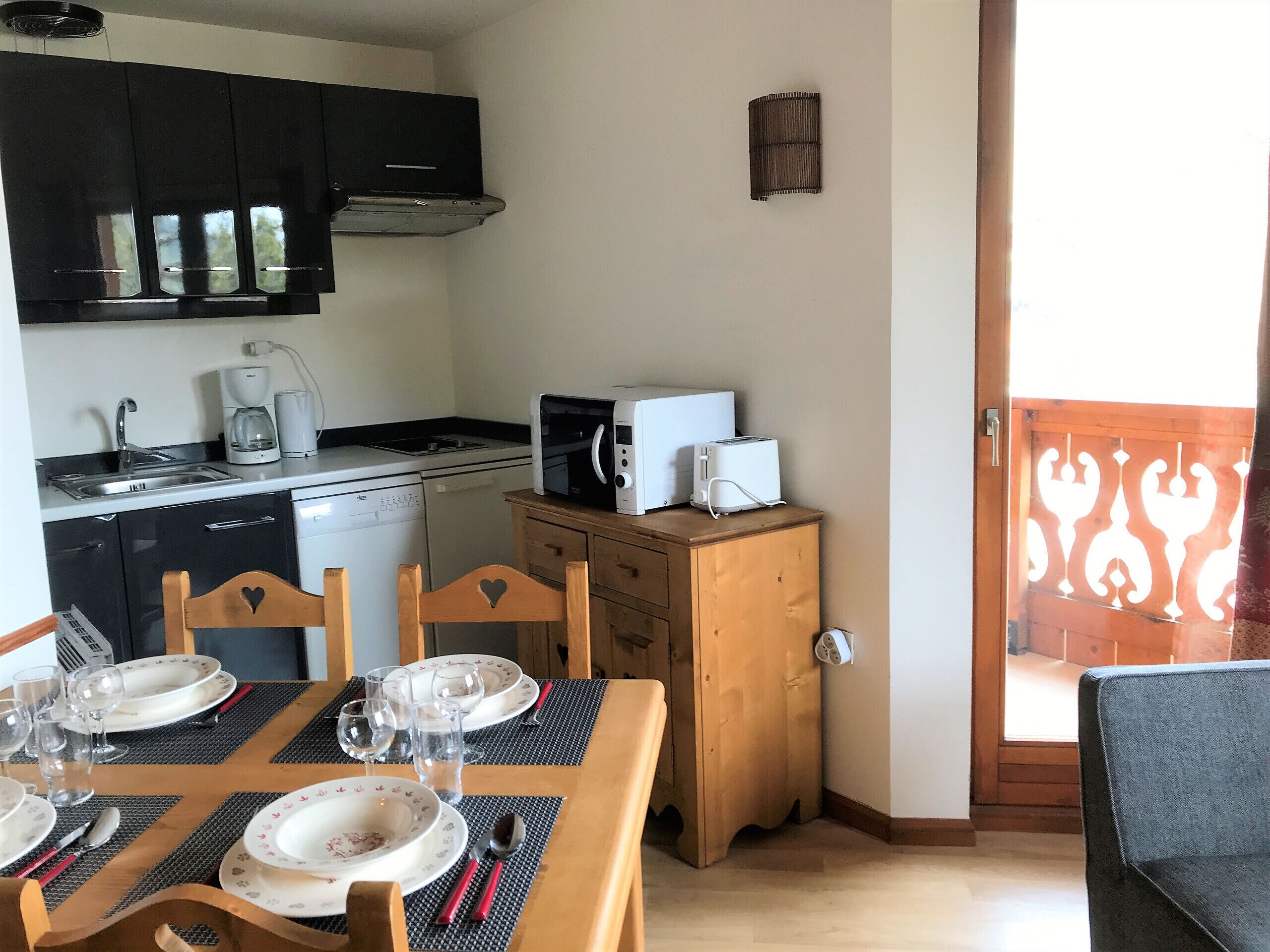 A compact kitchen with a dishwasher, microwave, and toaster, a dining table set with plates and cutlery, and a wooden cabinet with a heart design.