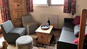 A cozy living room with a sofa, a chair, a small table with a bottle and glasses, a television on a wooden cabinet, and patterned curtains.