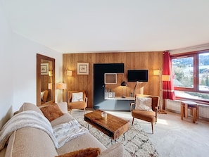 Ein Wohnzimmer mit einer Couch, Sesseln, einem Couchtisch, einem Fernseher und einem Schreibtisch mit einem Computer.