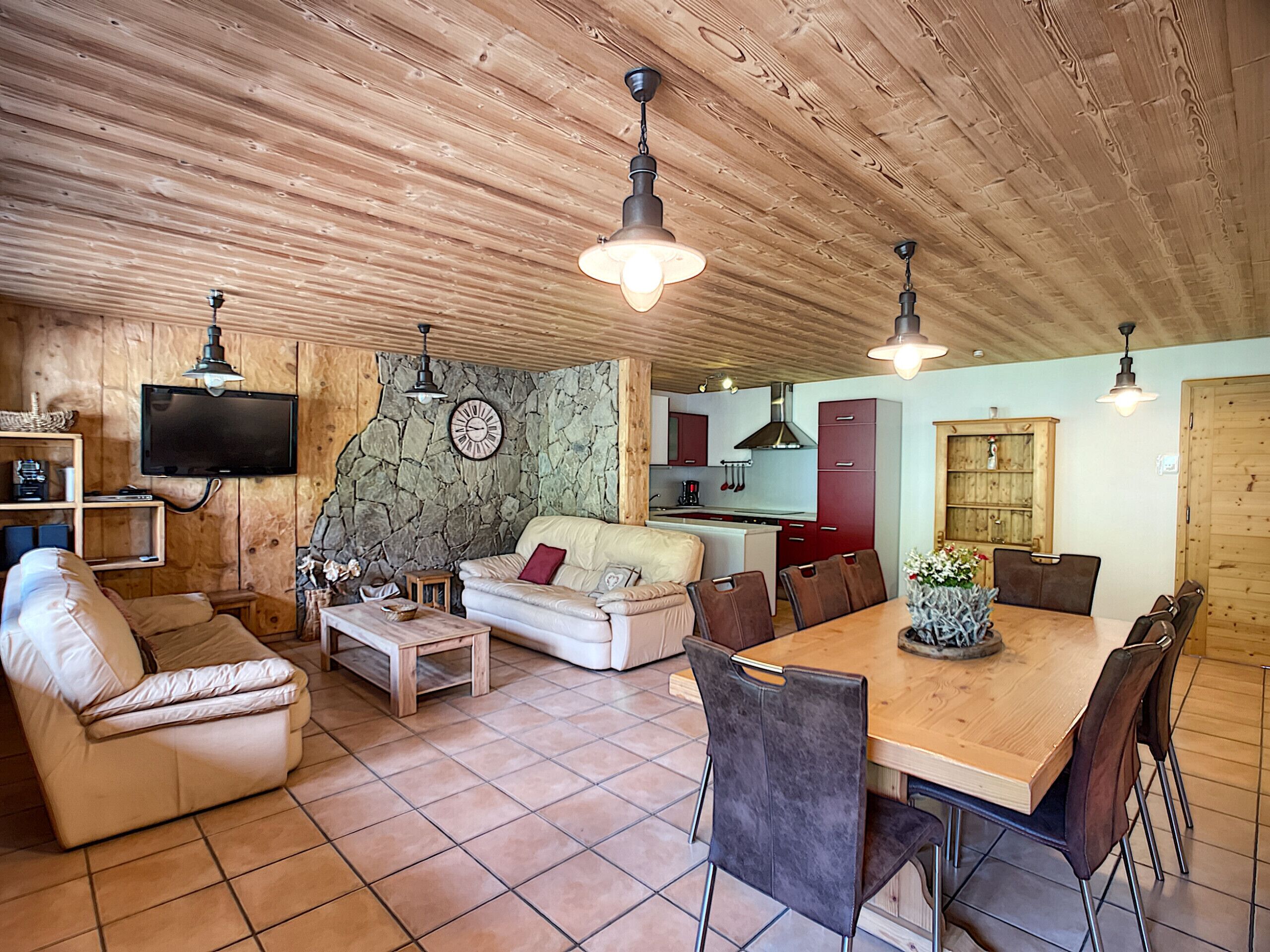 Uma sala de estar acolhedora, com um sofá, mesa de centro e mesa de jantar. É visível, ao fundo, uma cozinha com armários e uma parede de pedra.