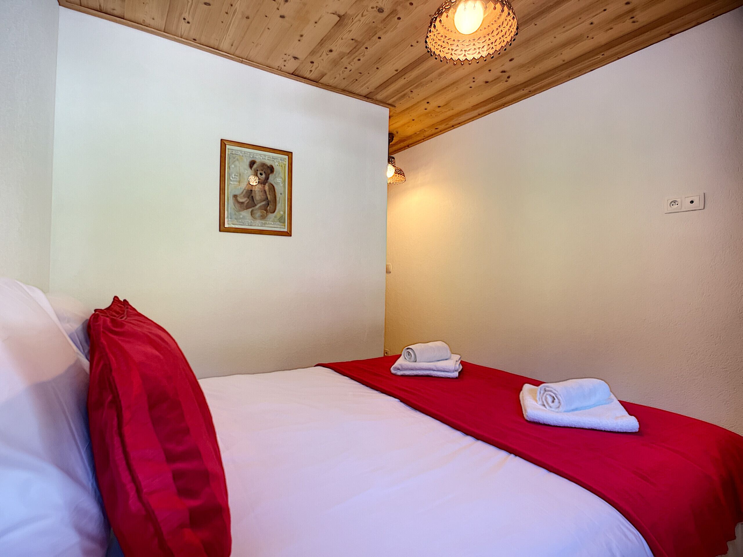 A hotel room with a wooden ceiling, a bed with red and white bedding, a framed picture on the wall, and a small lamp.