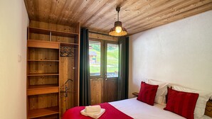 A bedroom with a wooden ceiling, a bed with red and white bedding, a wooden wardrobe, and a window with a view of greenery. - Koa - appartement - 8 pers - ski-in/out (Les Gets)