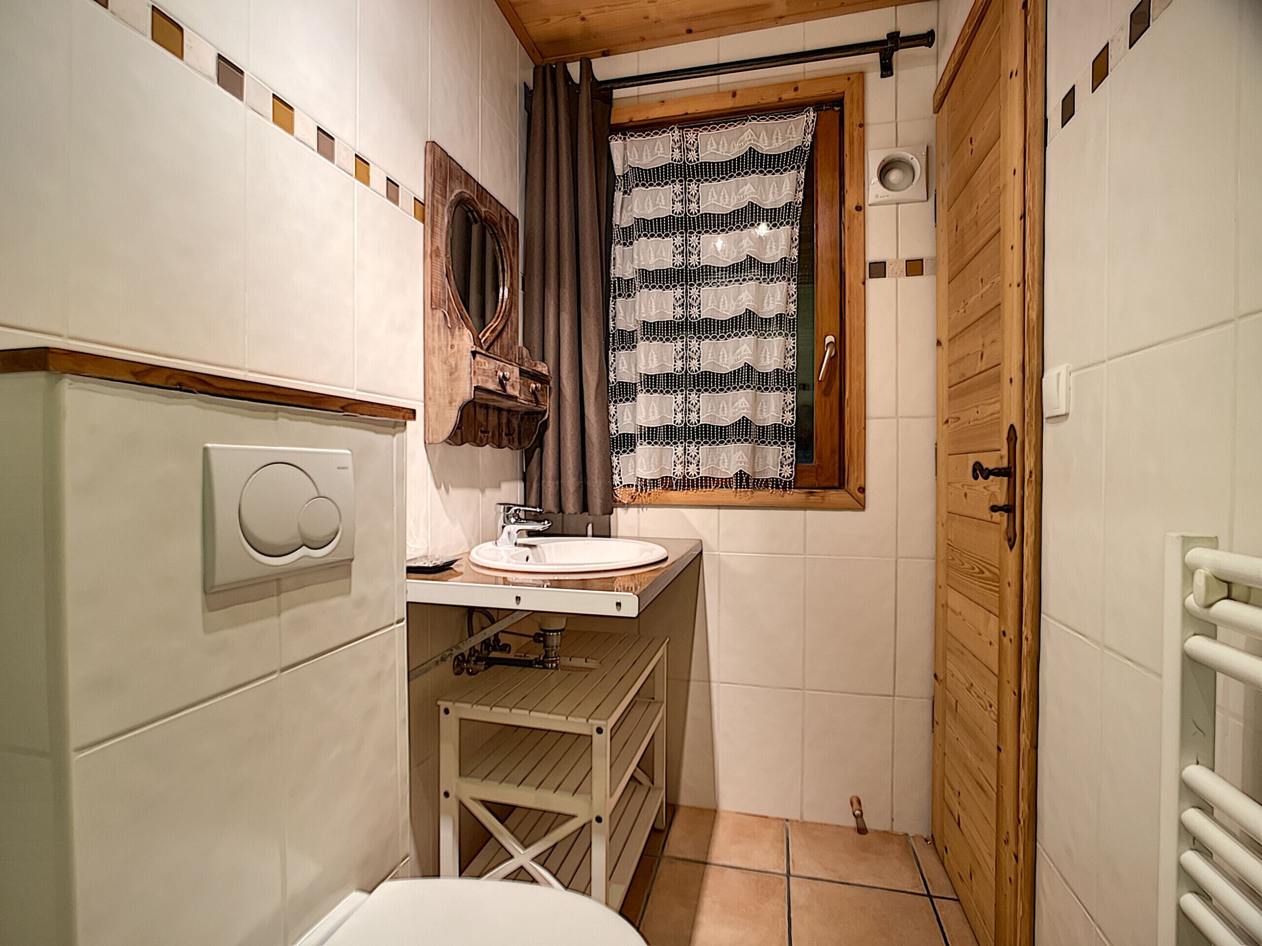 A compact bathroom with a wooden door, a white sink, a mirror, and a window with a patterned curtain.