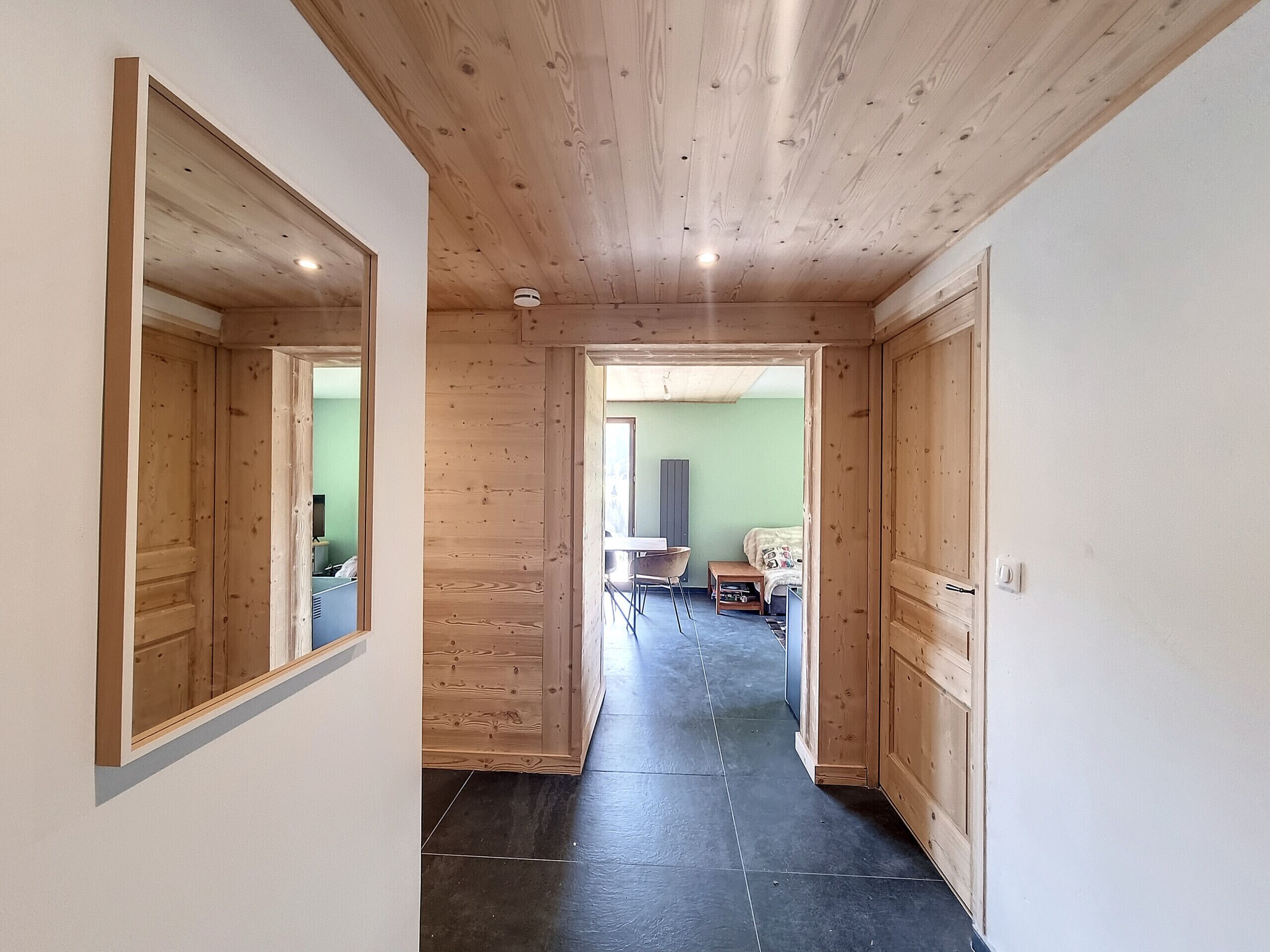 A wooden hallway with a mirrored wall, a door leading to a room with a bed and a TV, and a tiled floor.
