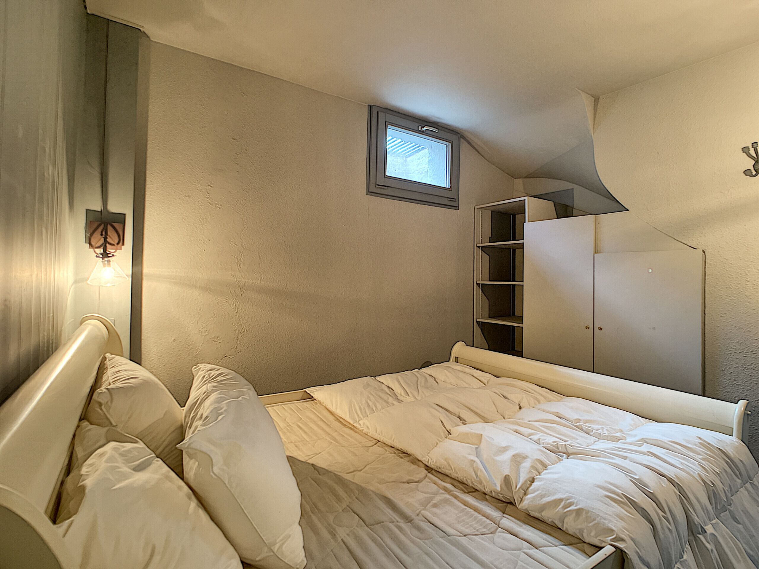 Une chambre avec un lit simple, une armoire, une petite fenêtre et un luminaire fixé au mur.