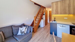A cozy living room with a grey sofa, wooden staircase, and a kitchen with wooden cabinets and a dishwasher.