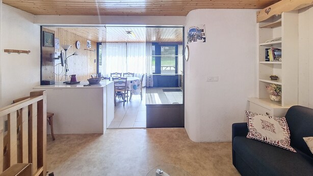 Une pièce avec un plafond en bois, un coin repas avec une table et des chaises, un canapé agrémenté d’un coussin à motifs et une bibliothèque.