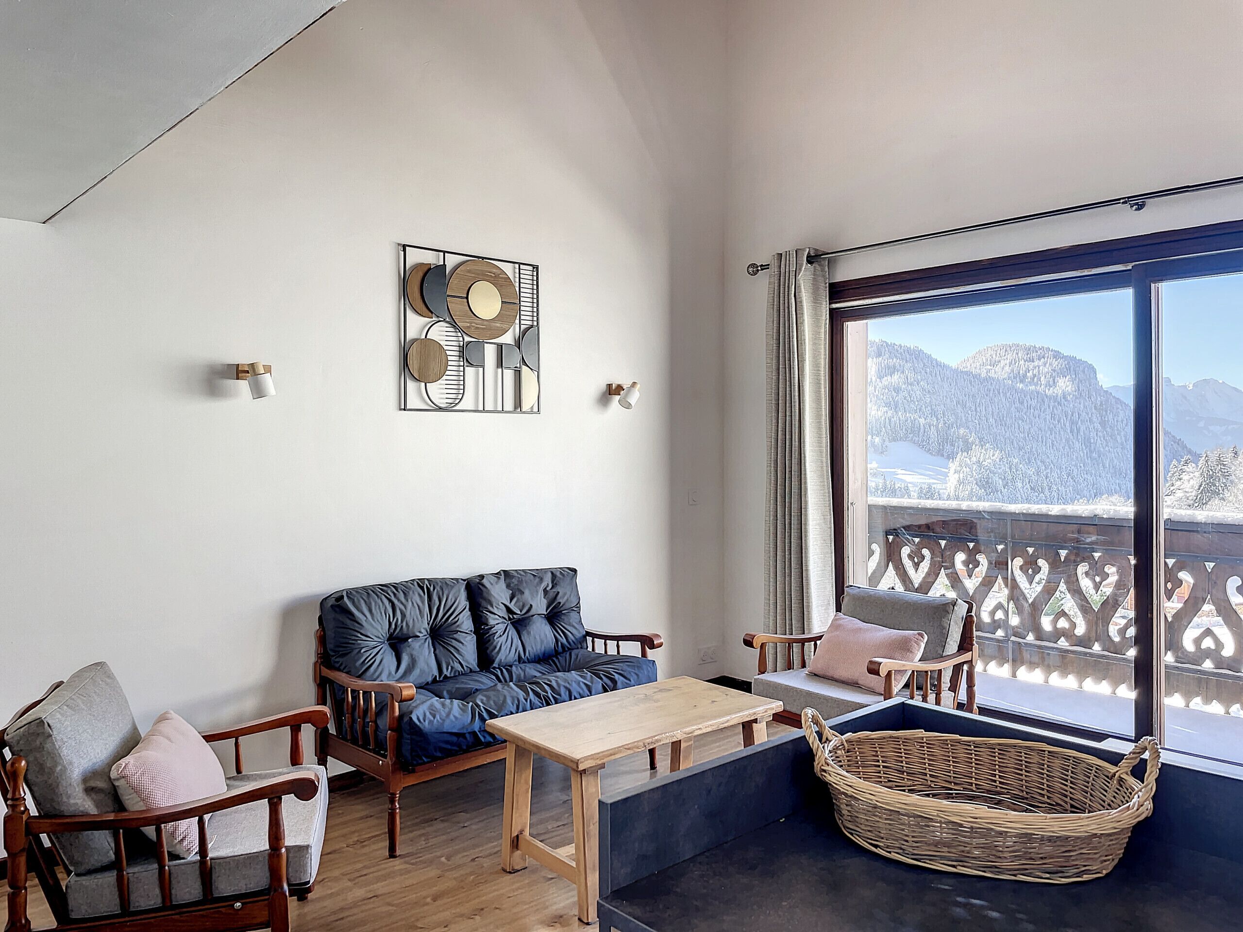 A cozy room with a blue sofa, wooden chairs, a small table, and a wicker basket. A large window offers a view of snow-covered mountains.